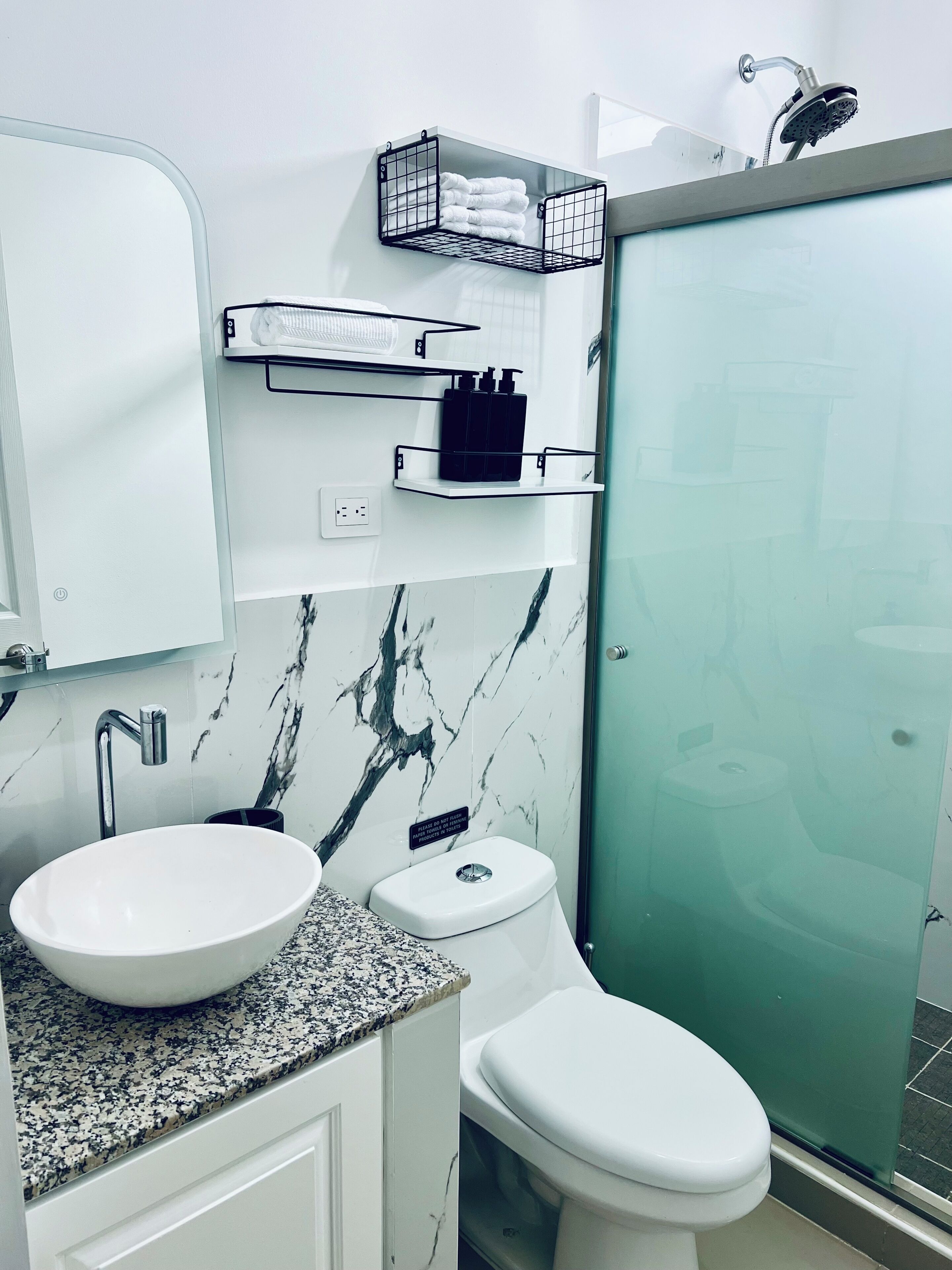 Un baño con lavamanos blanco, inodoro, cabina de ducha de vidrio y salpicadero de mármol.