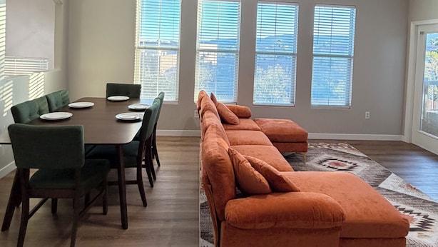 En spisestue med et bord dækket til seks personer, grønne polstrede stole, en stor orange modulsofa og en loftsvifte.