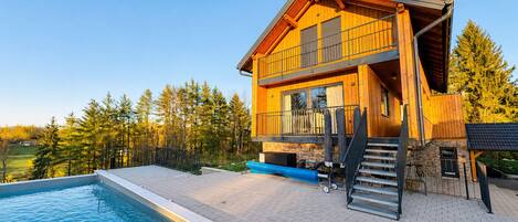 A two-story wooden house with a swimming pool, outdoor stairs, and a view of a forested area.
