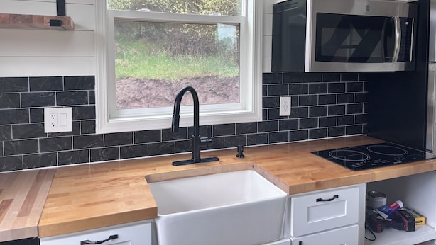 Una cocina con ventana, salpicadero de azulejos negros, armarios blancos, un microondas de acero inoxidable y una encimera de madera.