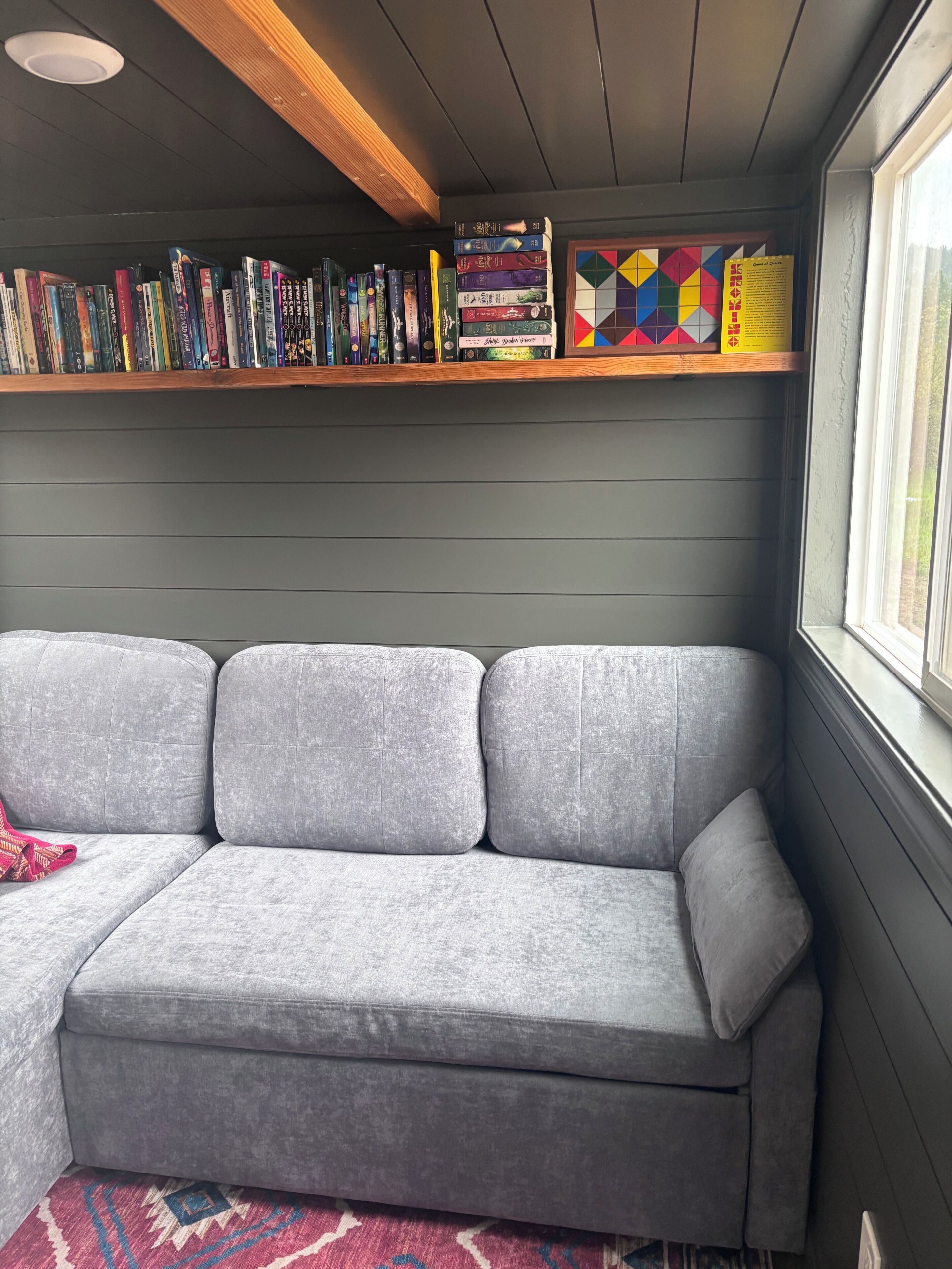 Ein graues Sofa mit einem gemusterten Teppich, ein Bücherregal mit Büchern und einer farbenfrohen, geometrischen Kiste, und ein Fenster mit Blick.