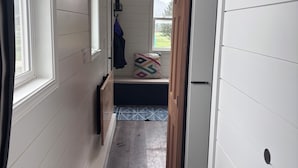 Un pasillo estrecho con paredes blancas y suelo de madera que conduce a un dormitorio con cama y ventana.