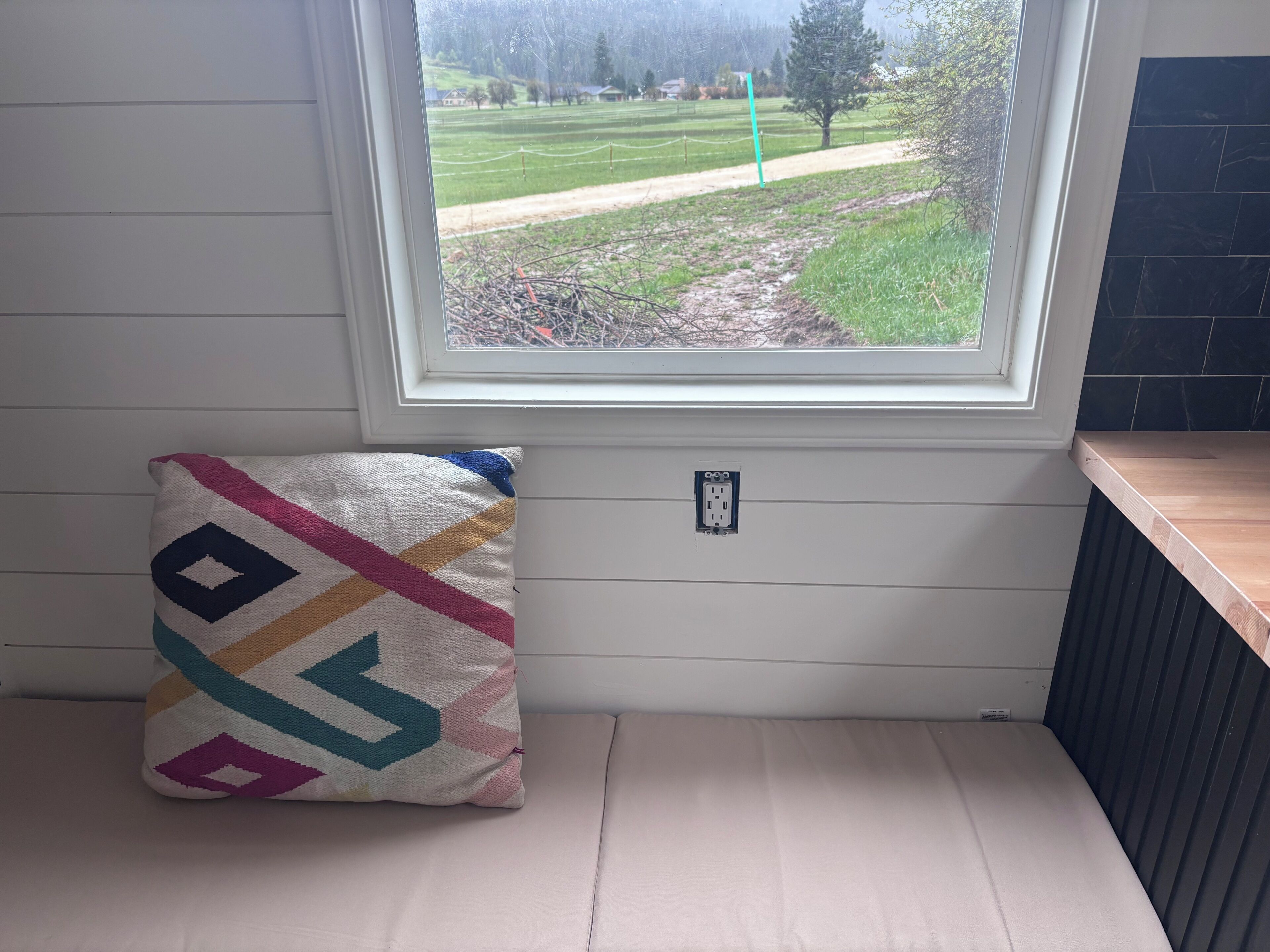 Ein Fenster mit Blick auf ein grasbewachsenes Feld und einen Baum, eine weiße Wand, eine Steckdose und ein farbenfrohes Kissen auf einer Bank.