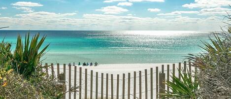 A beach with a wooden fence, clear turquoise water, and a few people sitting on the sand.