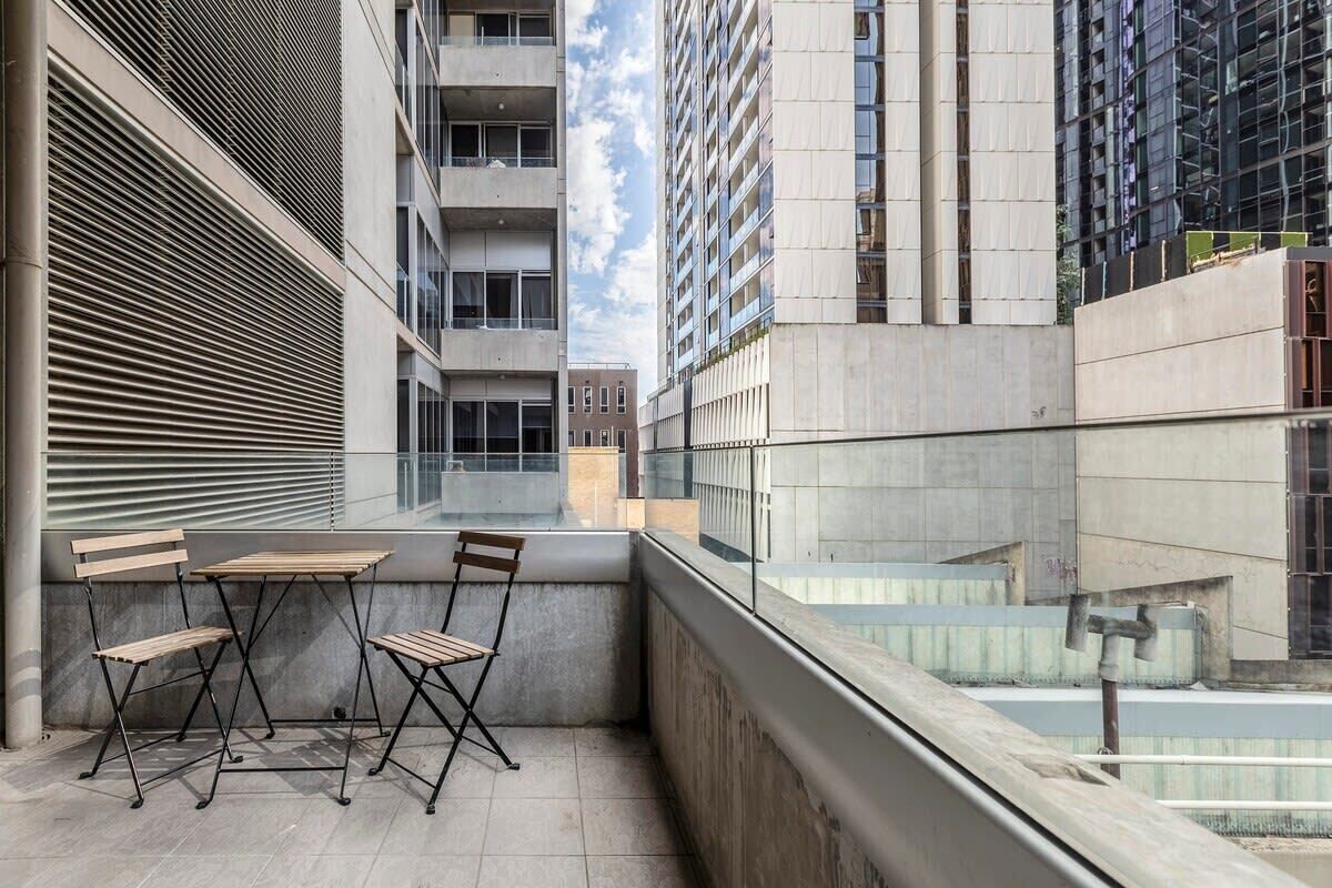 A balcony with a table and chairs, a view of adjacent buildings, and a clear sky.