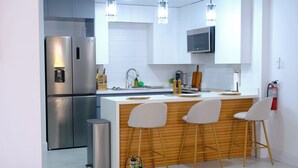 Una cocina moderna con electrodomésticos de acero inoxidable, salpicadero de azulejos blancos, taburetes de bar de madera e iluminación colgante.