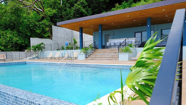Una piscina con borde de azulejos, un muelle de madera y una zona de estar cubierta con sillas y una escalera.