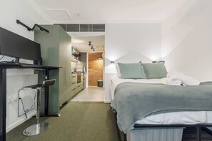 A compact hotel room with a bed, a desk, a chair, a lamp, and a kitchenette in the background. - Stylish studio apartment opposite Carlton Gardens (Carlton)