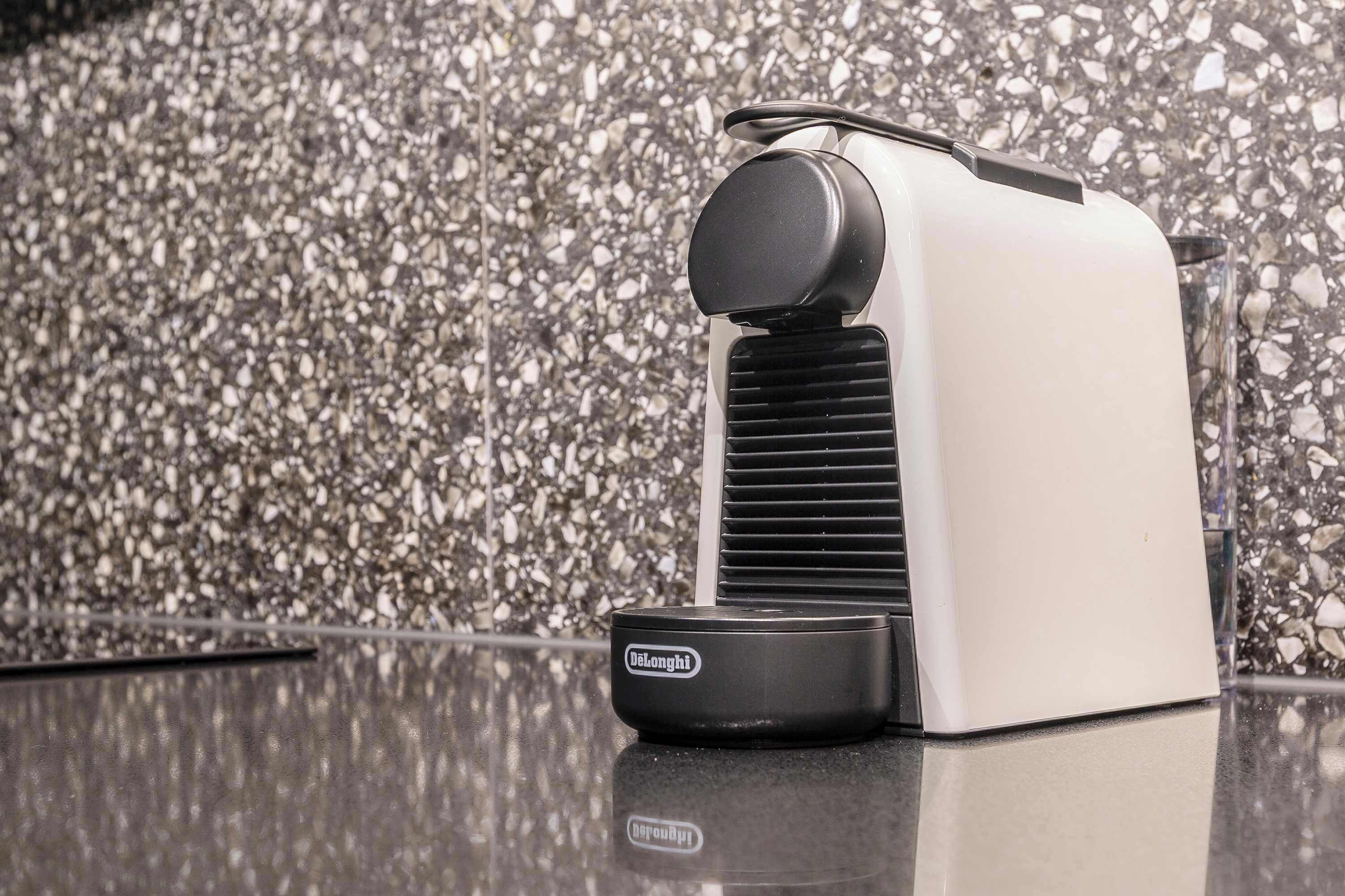A Delonghi espresso machine on a reflective surface with a terrazzo floor and a glass partition in the background.