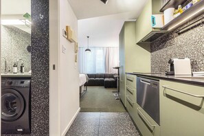 A compact kitchen with a washing machine, a dining area with a sofa, and a view of the outdoors. - Stylish studio apartment opposite Carlton Gardens (Carlton)