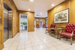 Hotellin aula, jossa on laatoitettu lattia, puupanelointi, kehystetty taideteos, punaisella kankaalla verhoillut tuolit ja lasiovi, joka johtaa ulos.