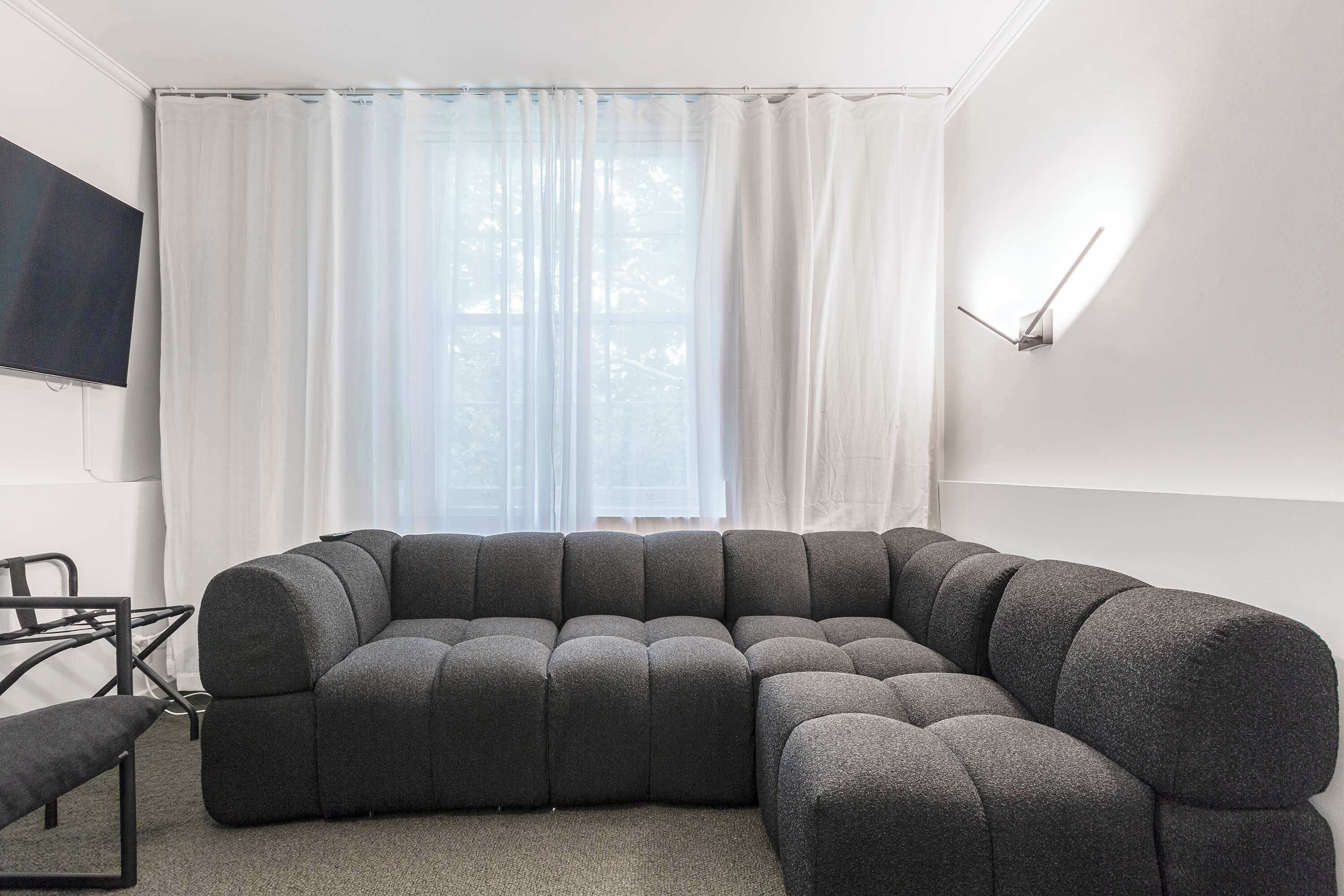A modern living room with a sectional sofa, a flat-screen TV mounted on the wall, and a large window with sheer curtains.