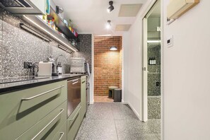 Uma cozinha com bancada de granito, armários verdes, parede de tijolos e uma pequena lixeira.