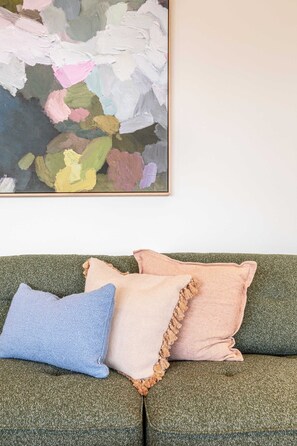 Ein grünes Sofa mit zwei Kissen, einem blauen und einem rosa, und einem farbenfrohen, abstrakten Gemälde an der Wand.