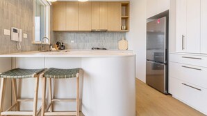 A modern kitchen with a central island, wooden stools, a stainless steel refrigerator, and white cabinetry. - Stylish, Spacious Apartment in Malvern (Malvern)