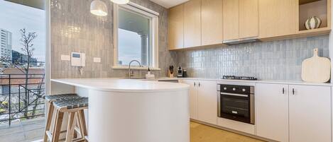 A modern kitchen with a central island, wooden stools, a large window, and a view of the city.