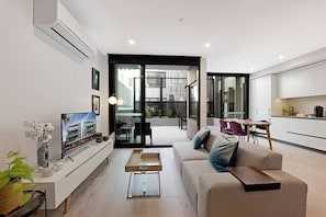 A modern living room with a sofa, TV, and a dining area with chairs and a table. - Stylish 1 Bedroom Apartment (North Melbourne)
