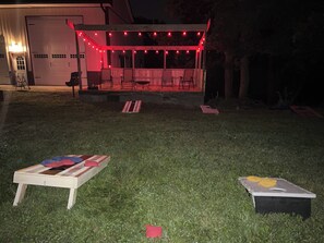 A covered outdoor seating area with string lights, a small table with a board game, and a grassy lawn.
