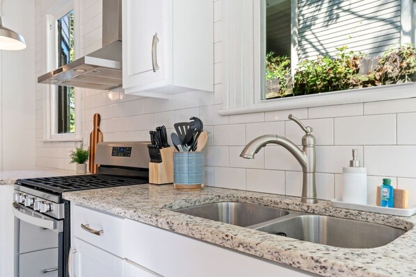A kitchen with white cabinets, a stainless steel sink, a stove, and a window with plants outside.