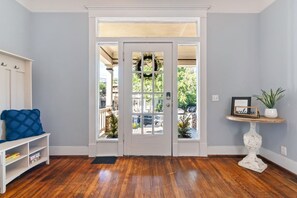 A room with a wooden floor, a white door with glass panels, a blue cushion, a small white shelf with a plant, and a welcome sign.