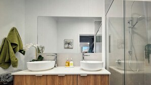 Ein modernes Badezimmer mit zwei Waschbecken, einem Holz-Badmöbel und einer Glasdusche. Ein Handtuchhalter mit einem grünen Handtuch ist ebenfalls vorhanden.