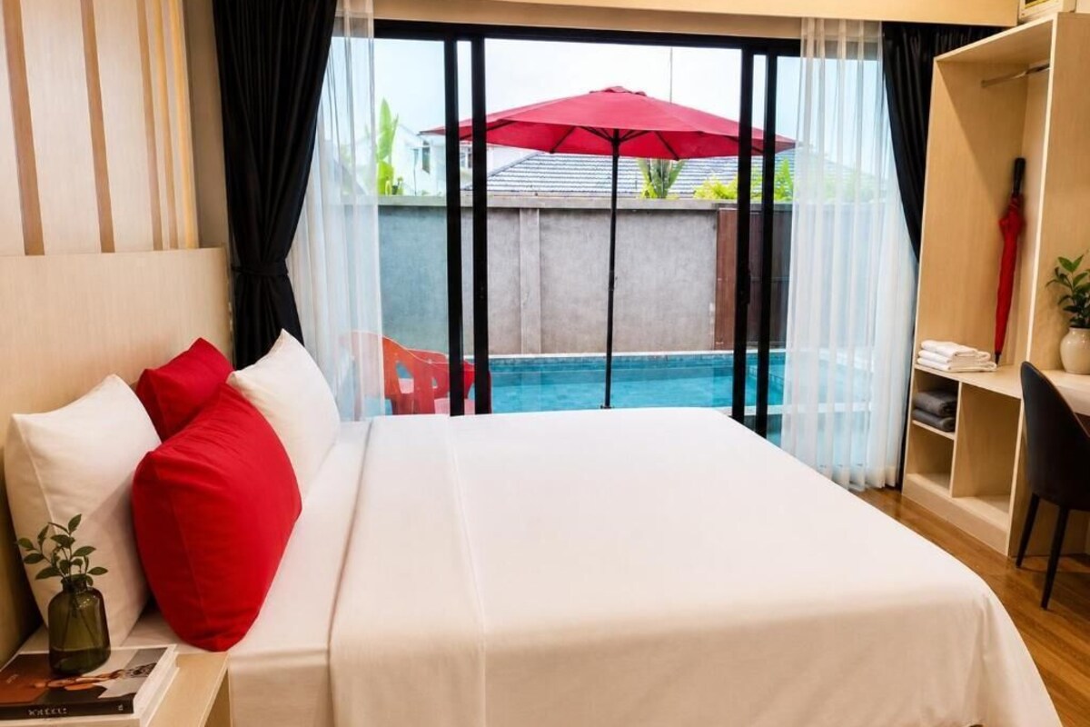 Habitación de hotel con cama, escritorio, silla, un paraguas rojo y vista a una piscina.