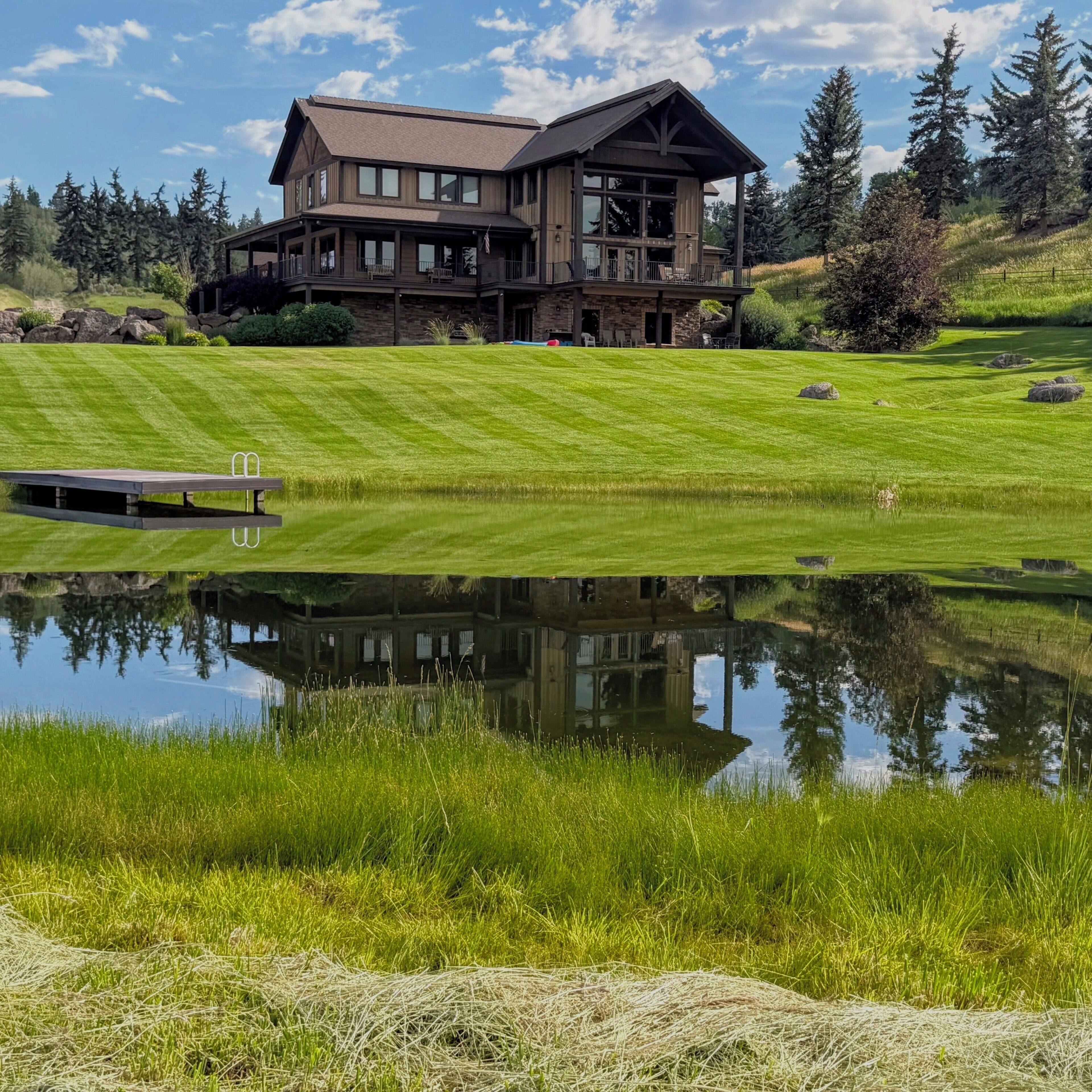 A large, two-story wooden house with a balcony, surrounded by a well-maintained lawn and a pond.
