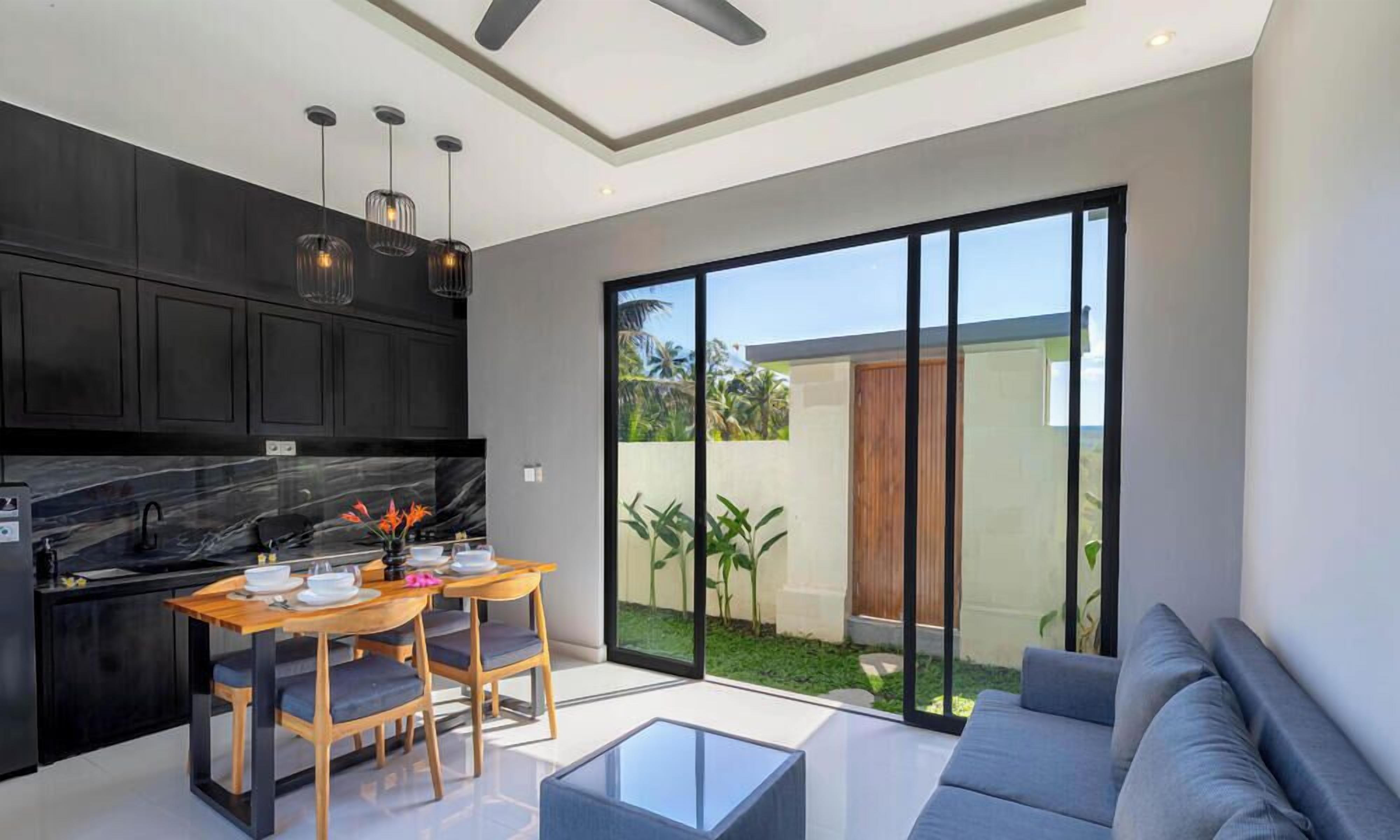 Cocina moderna con muebles oscuros, encimera de mármol e iluminación colgante. Cuenta con un comedor con mesa y sillas, y un amplio ventanal corredero que da a un jardín.