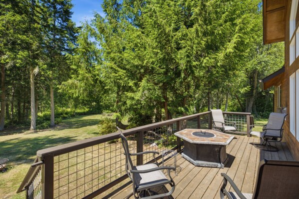A deck with outdoor furniture, a fire pit, and a view of a forested area. - Beachcombers Island Hideaway (Marrowstone)