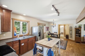 A kitchen with wooden cabinets, a stainless steel refrigerator, a sink, and a dining area with a table and chairs. - Beachcombers Island Hideaway (Marrowstone)
