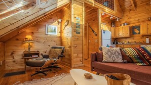 Ein gemütliches Blockhäuschen im Inneren mit Holzausstattung, einem Esstisch, einer Couch, einem Sessel, einer Lampe und einem Kühlschrank.