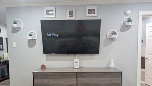 Una TV a schermo piatto montata a parete, con un mobiletto in legno sottostante, circondata da quadri incorniciati e piccoli scaffali con piante.