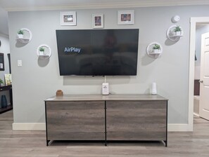 Ein Flachbildfernseher an einer Wand befestigt, darunter ein Holzkasten, umgeben von gerahmten Bildern und kleinen Regalen mit Pflanzen.