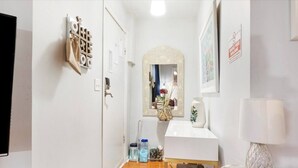 A hallway with a mirror, a small stool, a blue mat, a white shelf with a vase, and a wall-mounted lamp.