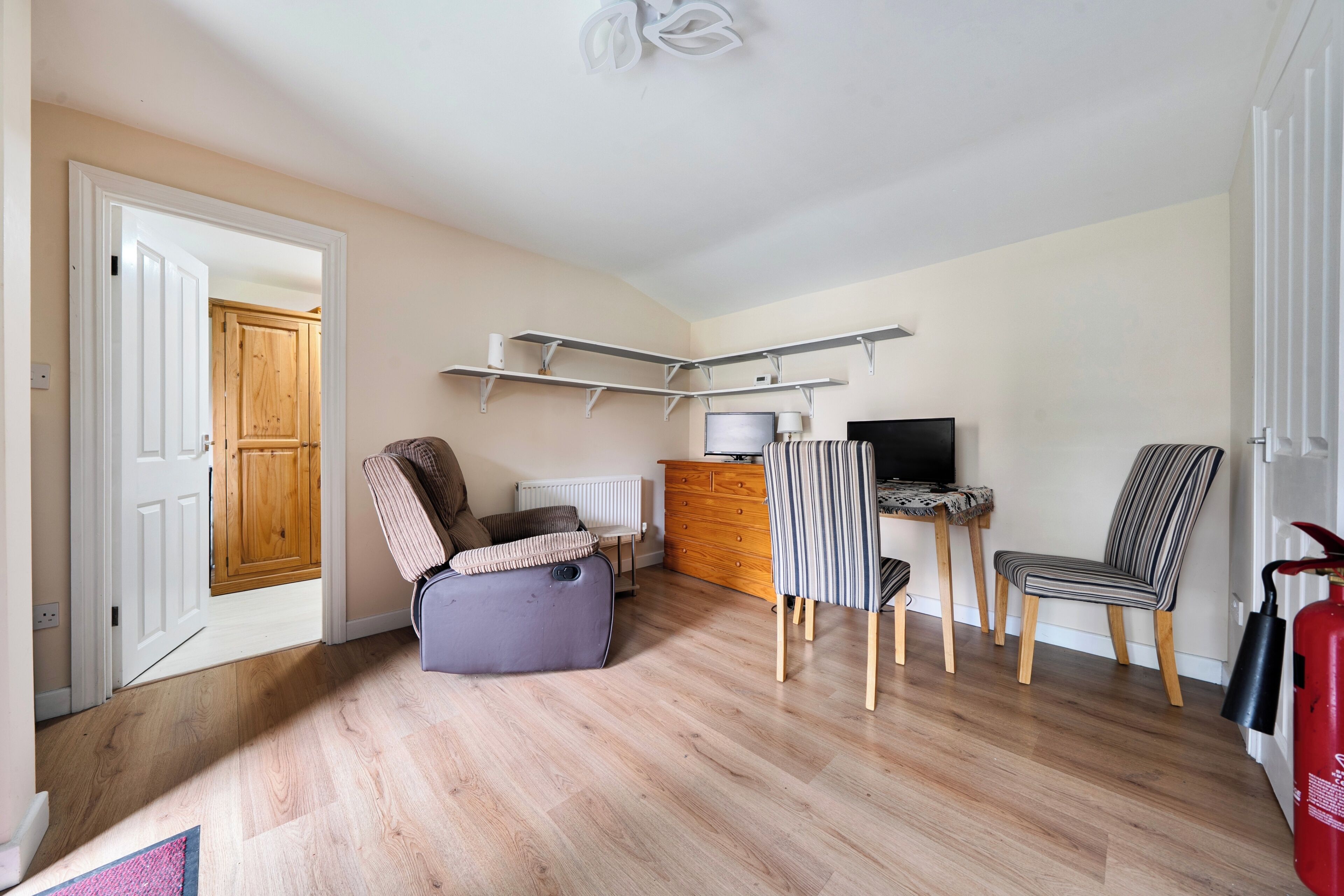 A room with a wooden floor, a desk with a computer, a chair, a small sofa, a radiator, and a fire extinguisher.