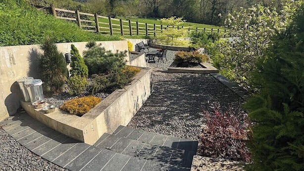 Anlagt udendørsområde med siddeplads, en betonplanter med beplantning og en grussti.