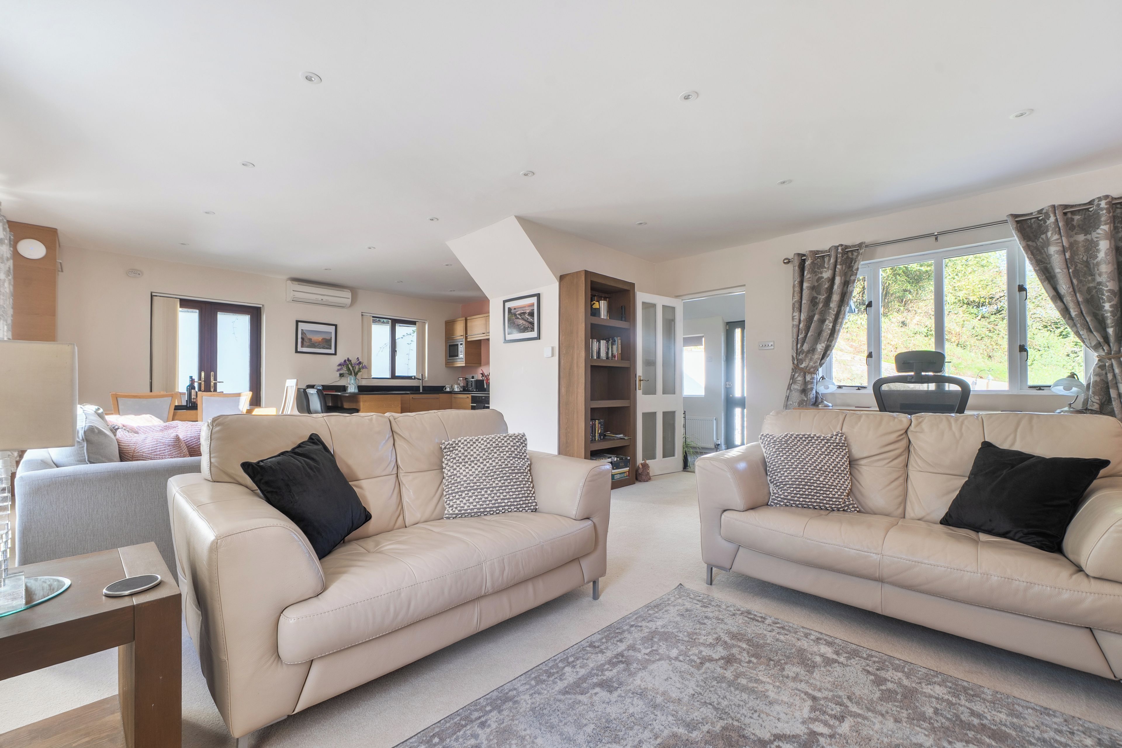 A spacious living area with two beige sofas, a coffee table, and a rug. There is a kitchen in the background with cabinets and a dining area.