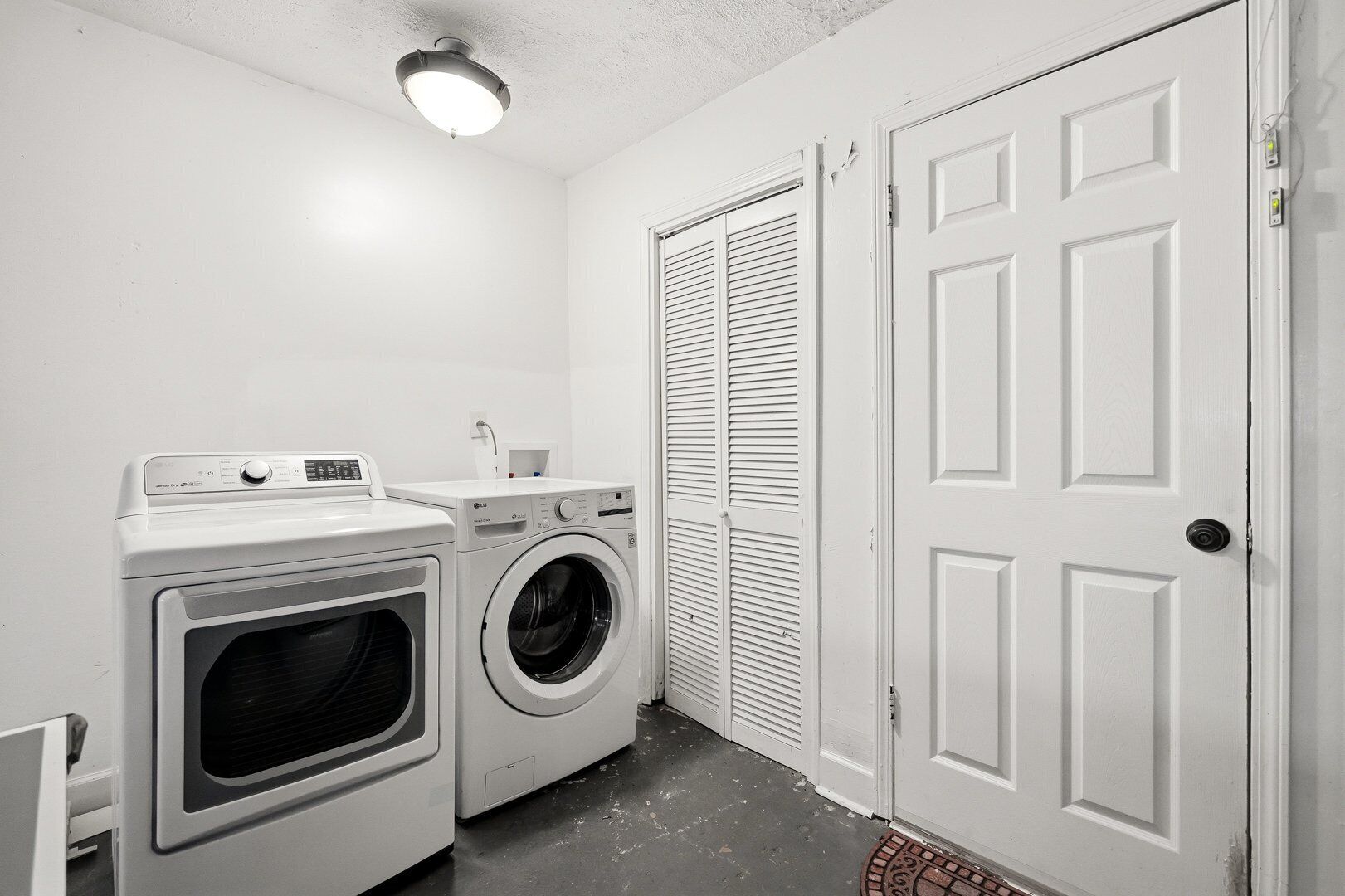 A laundry room with two washing machines, a dryer, a ceiling light, and a white door.
