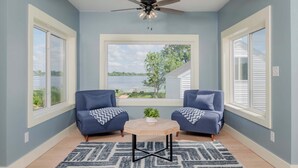 Ein Zimmer mit Deckenventilator, zwei blaue Sessel, einem kleinen Tisch mit Pflanze und Seeblick durch ein großes Fenster.