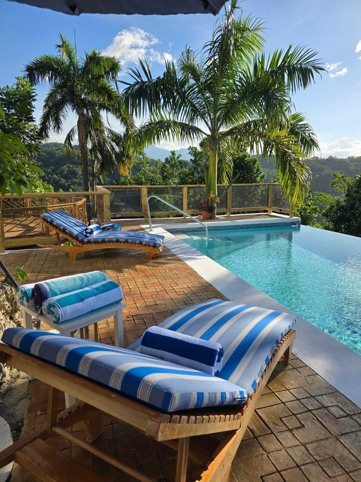 A poolside area with lounge chairs and a view of palm trees and hills.
