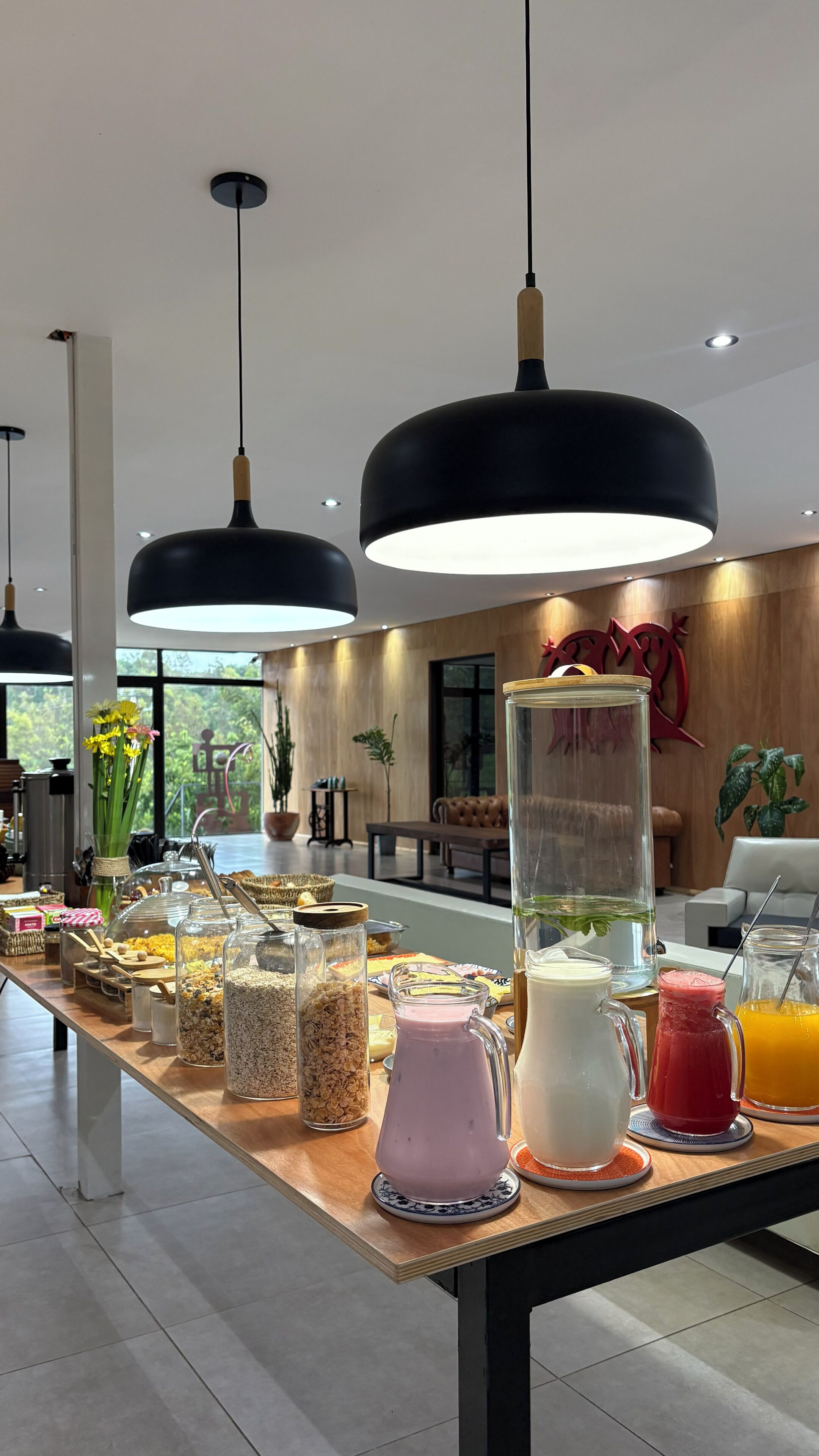 A breakfast buffet with various food items and drinks, modern pendant lighting, and a view of the outdoors.