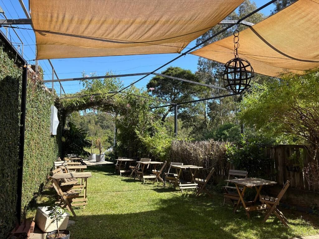 An outdoor patio with tables and chairs under a shaded area, surrounded by greenery.