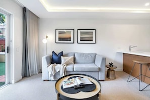 A modern living room with a grey sofa, a coffee table with a magazine, two framed pictures on the wall, and a kitchen area with a bar stool.