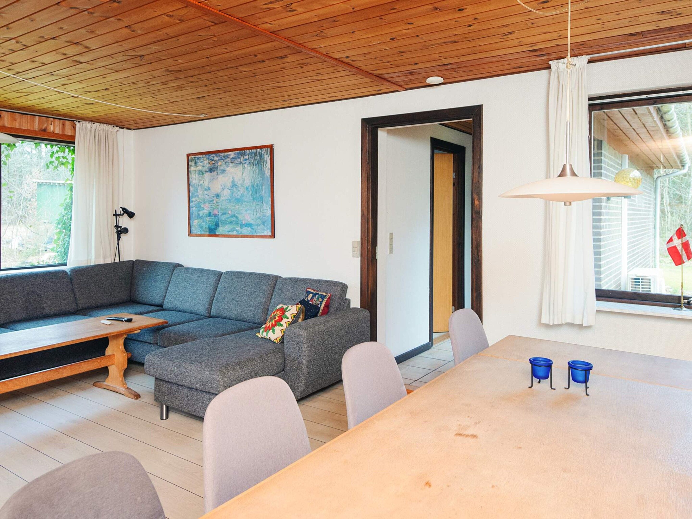 A living room with a grey sectional sofa, a wooden coffee table, a dining table with chairs, a painting on the wall, and a window with curtains.