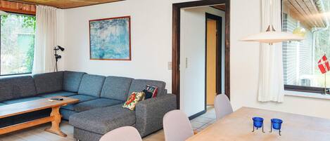A living room with a grey sectional sofa, a wooden coffee table, a dining table with chairs, a painting on the wall, and a window with curtains.