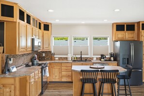 A kitchen with wooden cabinets, a central island with bar stools, a refrigerator, and a view of a lake through windows. - Gateway to Paradise | Rainier Views in Packwood (Packwood)