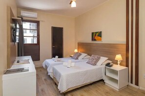 Ein Hotelzimmer mit zwei Betten, einem Schreibtisch, einem Stuhl, einem Nachttisch mit Lampe, einer Wandklimaanlage und einer Tür.