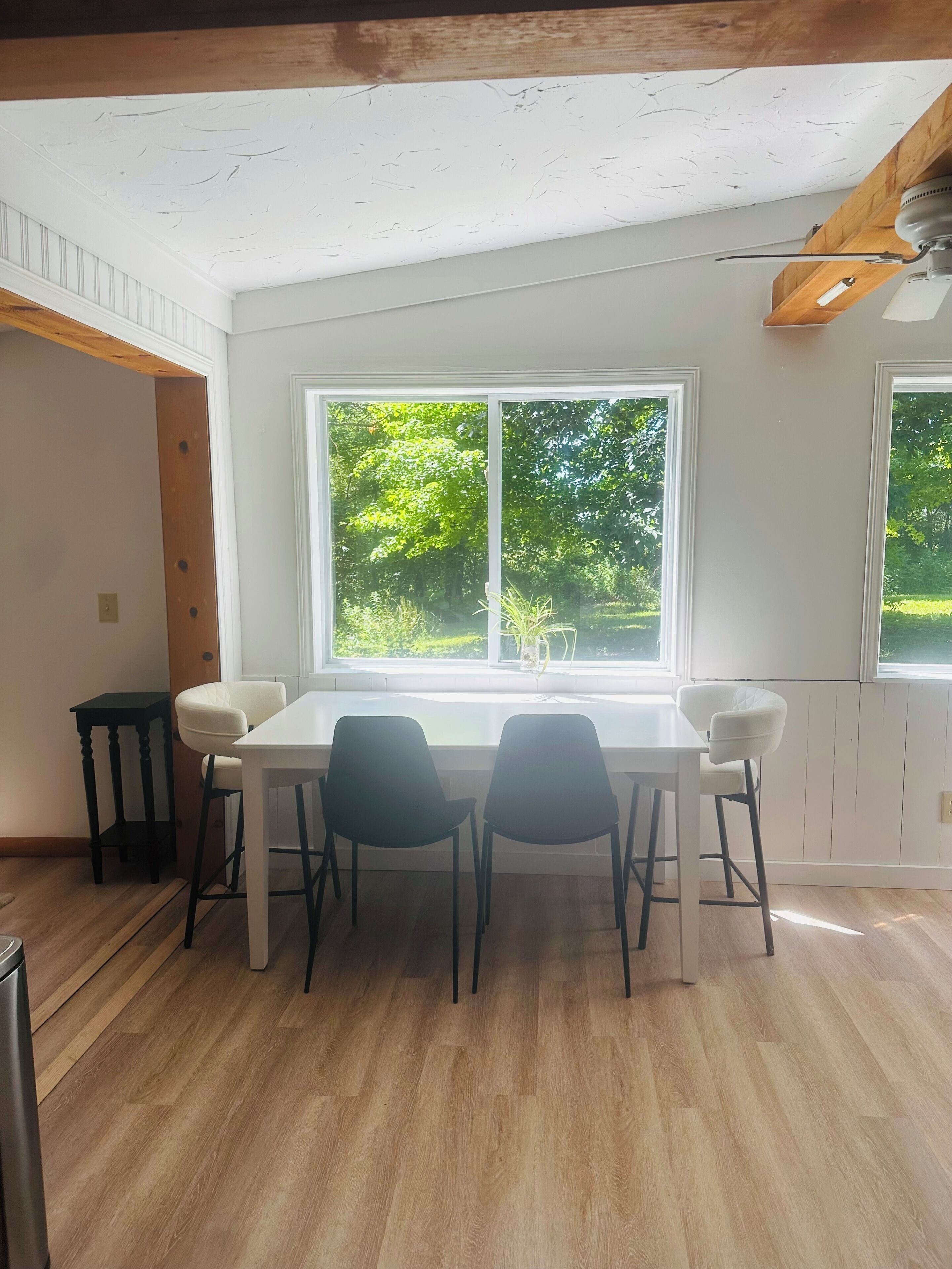 Un coin repas avec une table blanche et des chaises noires, de grandes fenêtres donnant sur un paysage verdoyant et un ventilateur de plafond.
