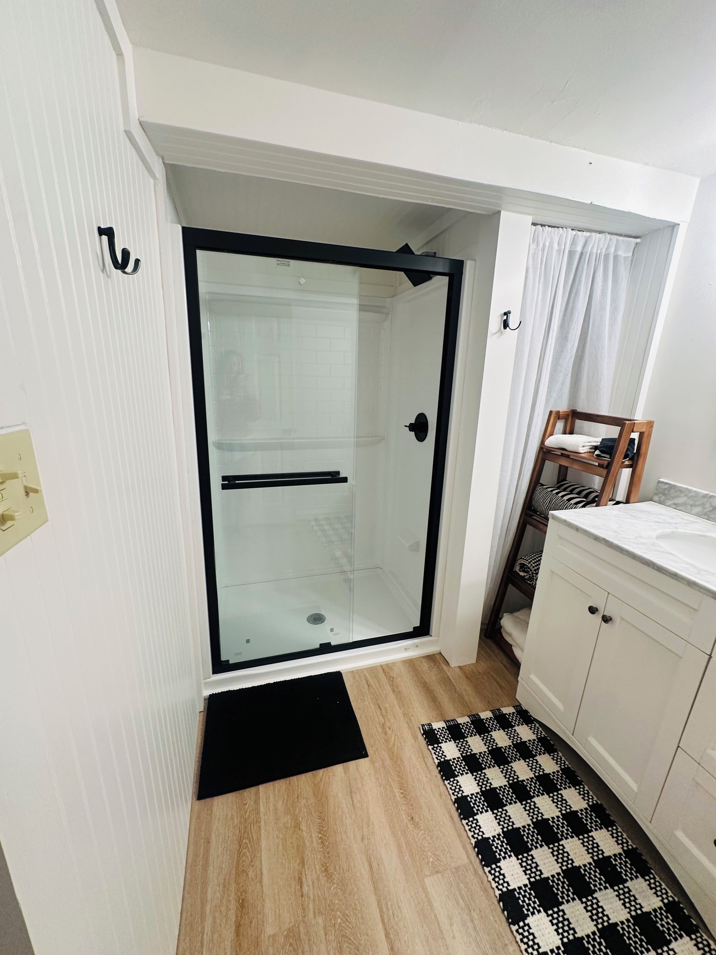 Un baño con cabina de ducha de vidrio, una silla de madera, un mueble blanco y una alfombra de cuadros.