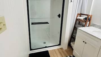 Un baño con cabina de ducha de vidrio, una silla de madera, un mueble blanco y una alfombra de cuadros.